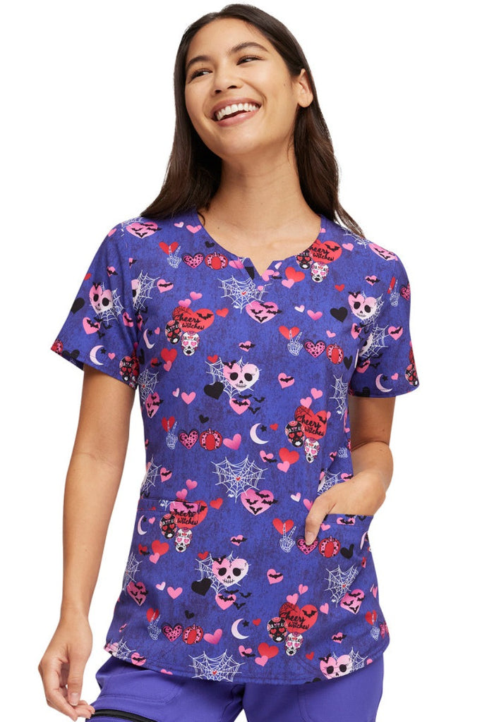 A young female Pediatric Nurse wearing a HeartSoul Women's V-Neck Print Scrub Top in "Cheers Witches" featuring spooky skulls and hearts with spiderwebs and bats scattered throughout the print.