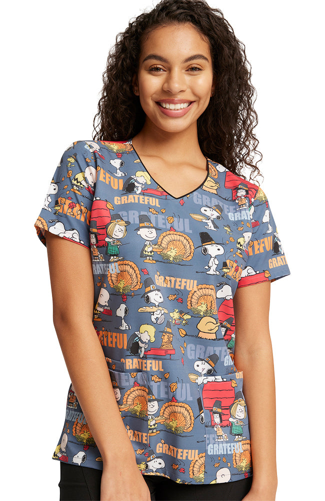 A young female Pediatric Nurse wearing a Tooniforms Women's Printed Scrub Top in "Grateful Snoopy" featuring the whole Peanuts Gang with Pilgrim Snoopy scattered throughout the print.