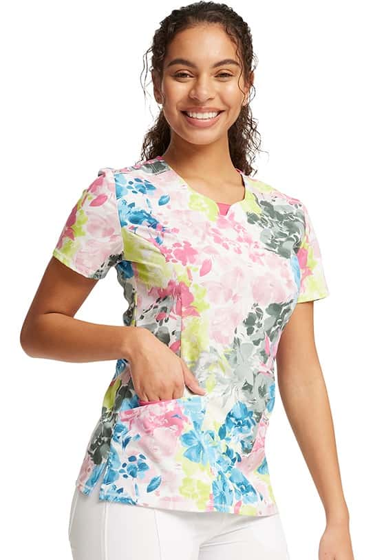 A female Certified Nurse's Assistant wearing a Cherokee Infinity Women's Round Neck Print Top in "Brush Away Blooms" featuring 2 front patch pockets with 1 interior pocket on the right side.