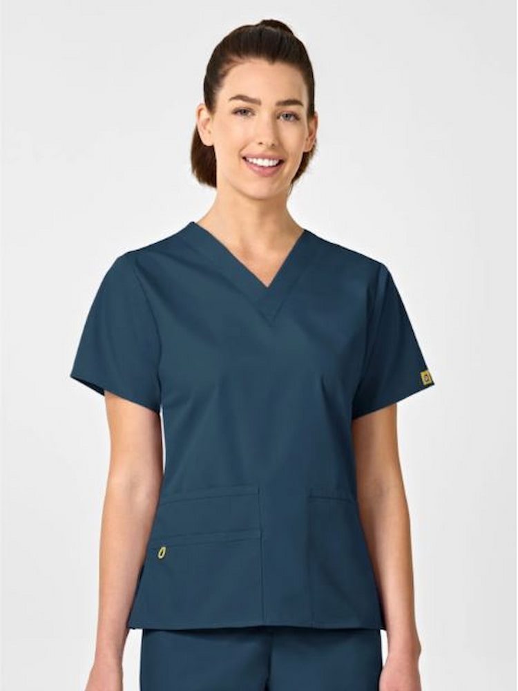 A young female Medical Assistant wearing a WonderWink Origins Women's Bravo Scrub Top in Caribbean Blue size Large featuring a soft poly cotton blended fabric.