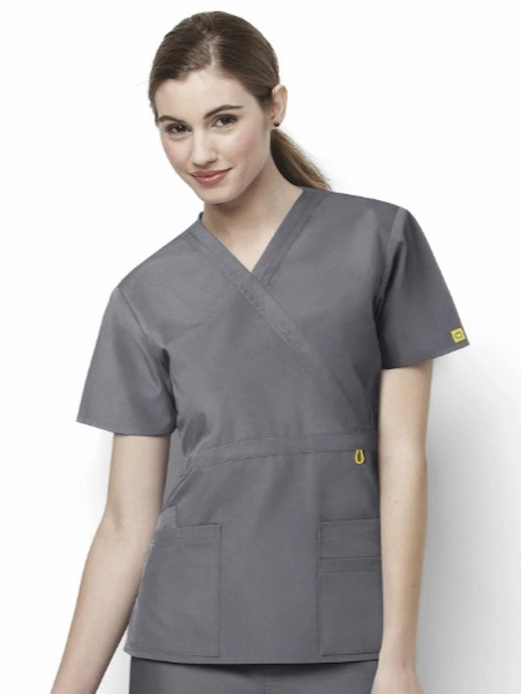 A young female LPN wearing a WonderWink Origins Women's Fashion Waist Scrub Top in Pewter size Large featuring a front mock belt with bust darts for shaping.