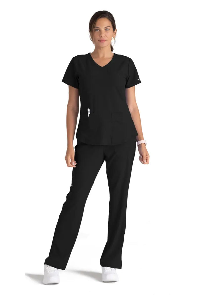 A full body image of a young female LPN wearing a Skechers Women's Breeze V-neck Scrub Top in Black size XL featuring two front patch pockets and a utility pocket on the wearer's left side.