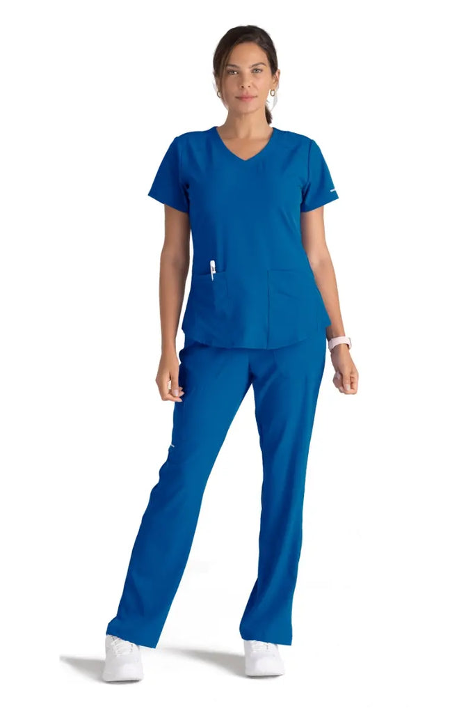 A full body image of a young female Surgeon wearing a Skechers Women's Breeze V-neck Scrub Top in Royal Blue size XL featuring two front patch pockets and a utility pocket on the wearer's left side.