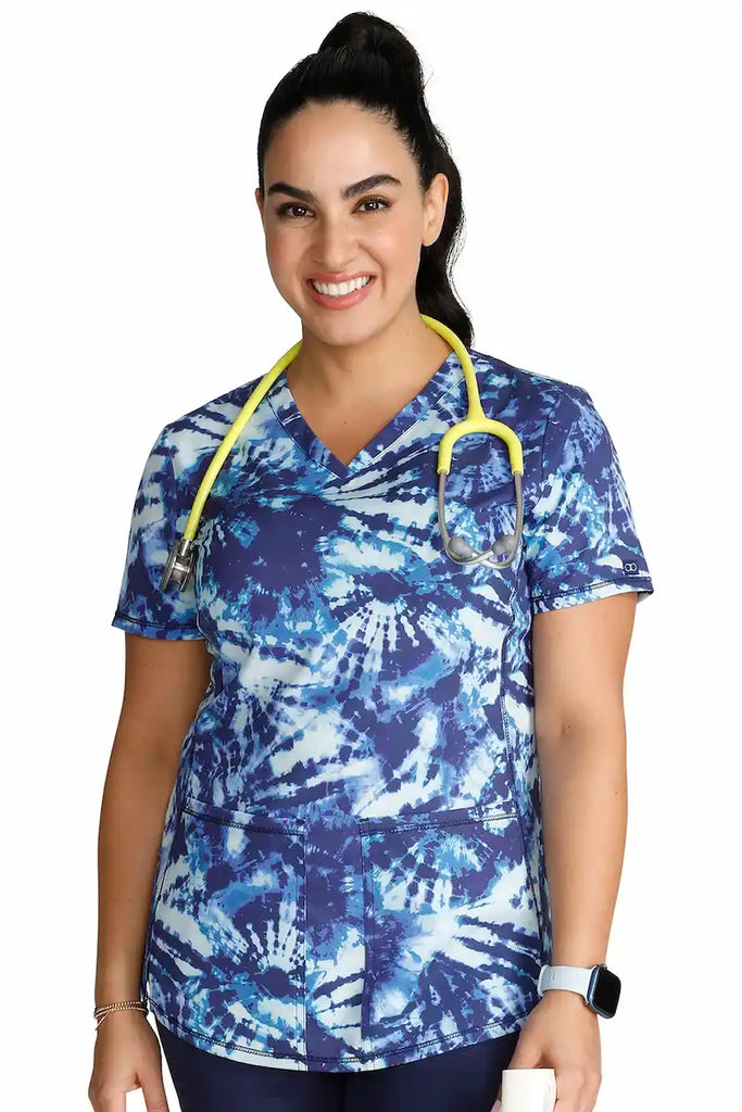 A young female Oncology Nurse wearing a Cherokee Women's V-neck Printed Scrub Top in "Tie Dye Tranquility" size XS featuring calming colors and swirling patterns that promote a sense of relaxation and well-being.