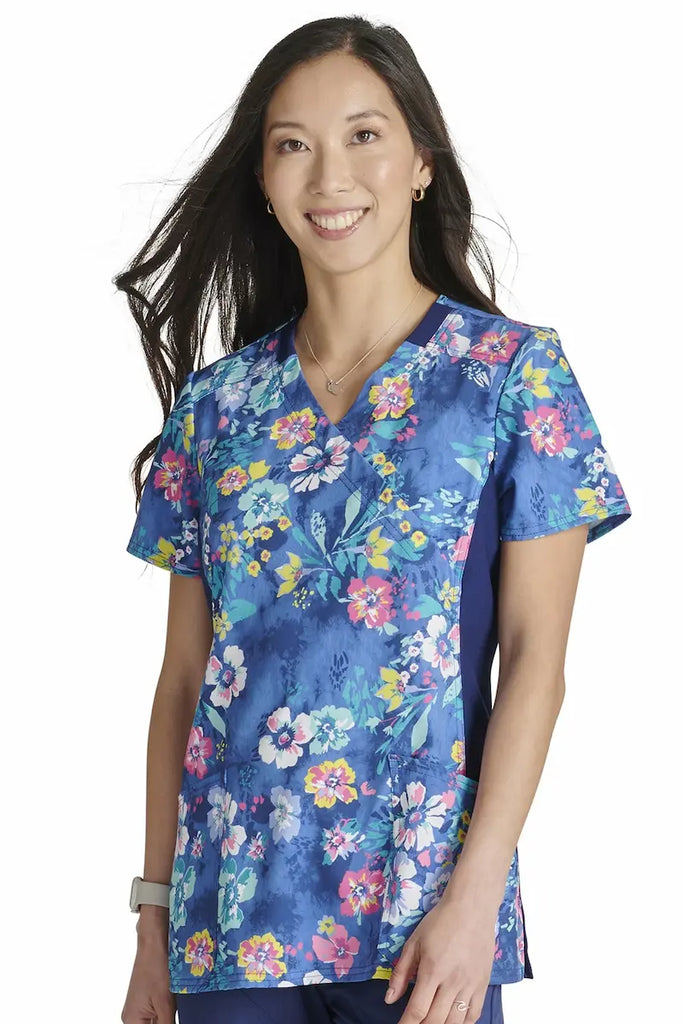 A young female Neonatal Nurse wearing a Cherokee Women's Knit Panel Printed Scrub Top in "Blooming Tie Dye" size XS featuring a stylish mock wrap neckline.