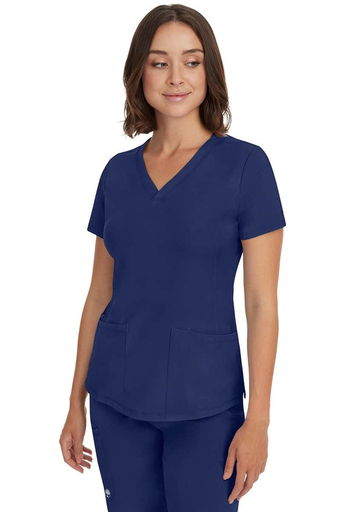 A young female healthcare professional wearing a HH-Works Women's Monica Multi-Pocket Scrub Top in Navy featuring a center back length of 24".