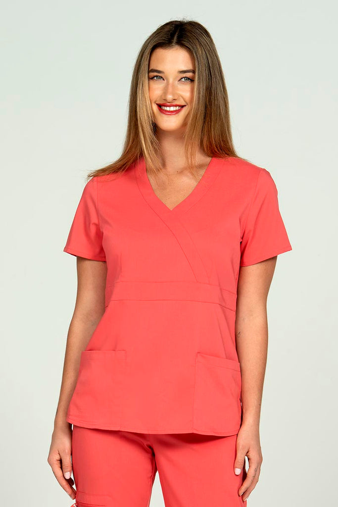 A young female CNA wearing an Epic by MedWorks Women's Mock Wrap Scrub Top in Coral size small featuring a stylish front seaming.