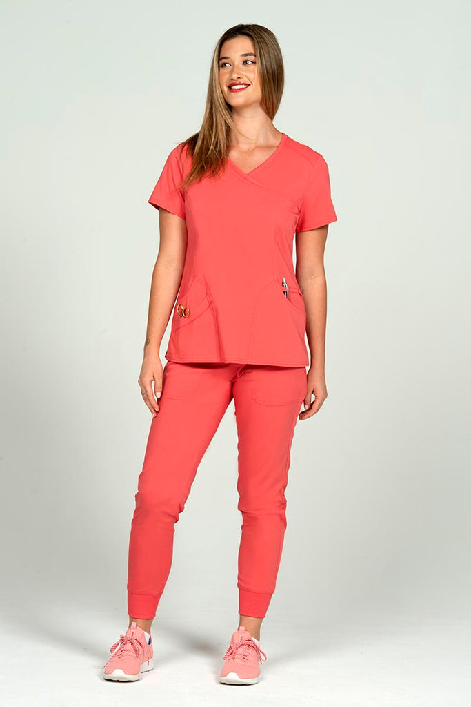A full body image of a young female Nurse Practitioner wearing an Epic by MedWorks Women's Knit Collar Mock Wrap Scrub Top in Coral size XL featuring a total of 4 pockets.