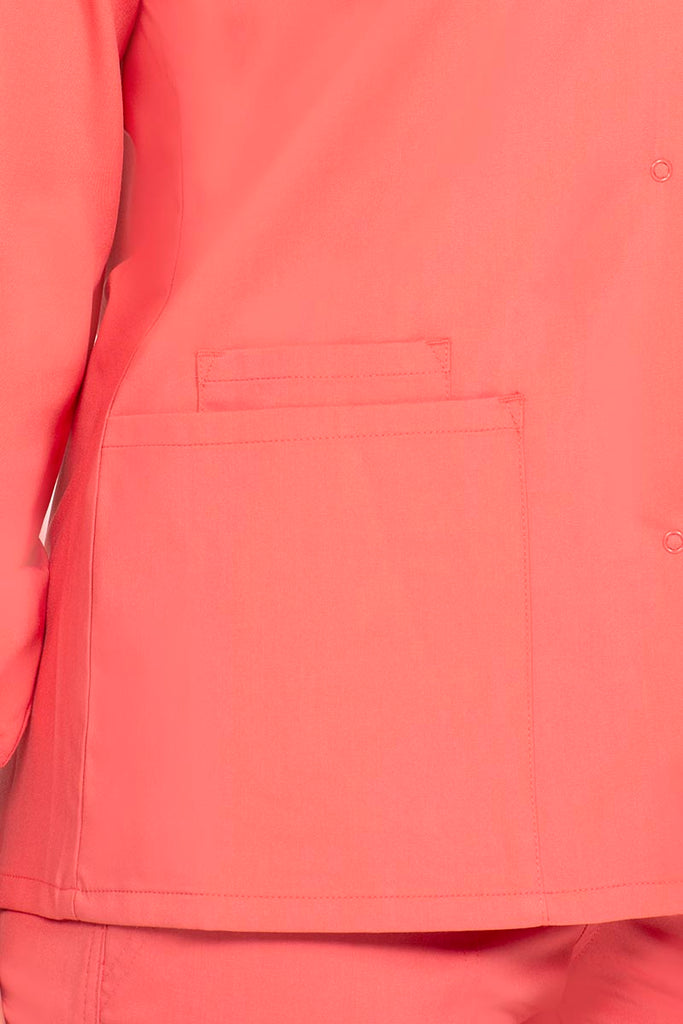 A young female RN wearing an Epic by MedWorks Women's Snap Front Scrub Jacket in Coral size Medium featuring 1 interior cellphone pocket pocket on the wearer's right side.