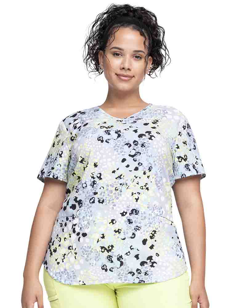 Young female healthcare worker wearing a Cherokee Women's V-Neck Print Scrub Top in "Spots Go" featuring a curved hemline.