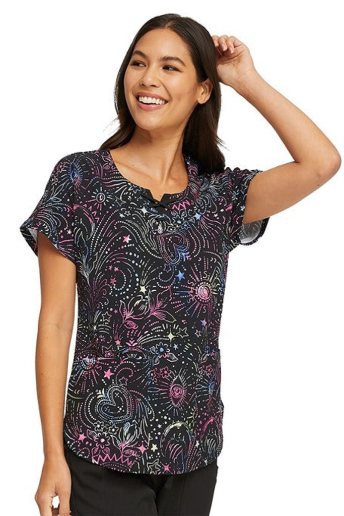 A young female Nurse Practitioner wearing a HeartSoul Women's Round Neck Print Scrub Top in "Celestial Twist" size Medium featuring 2 front patch pockets.