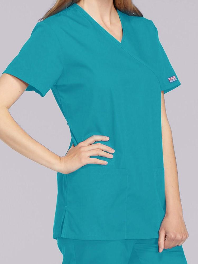 A young female Pharmacist wearing a Cherokee Workwear Originals Women's Mock Wrap Solid Scrub Top in Teal size Small featuring a 2 front patch pockets.