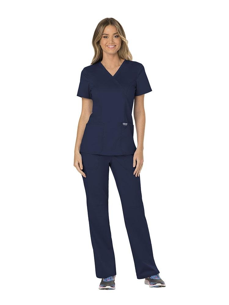 A young female Medical Assistant wearing a Cherokee Workwear Revolution Women's Mock Wrap Scrub Top in Navy size XXS featuring 2 front patch pockets.