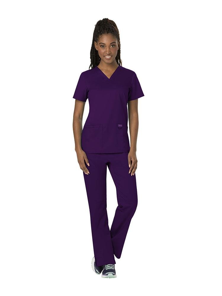 A young female Registered Nurse wearing both the Cherokee Workwear Revolution women's V-Neck Scrub Top & Elastic Waistband Pull-On Scrub Pant in Eggplant size extra small.