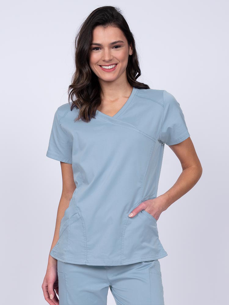 Young RN wearing an Epic by MedWorks Women's Knit Collar Mock Wrap Scrub Top in blue fog featuring a Y-neckline with short sleeves.