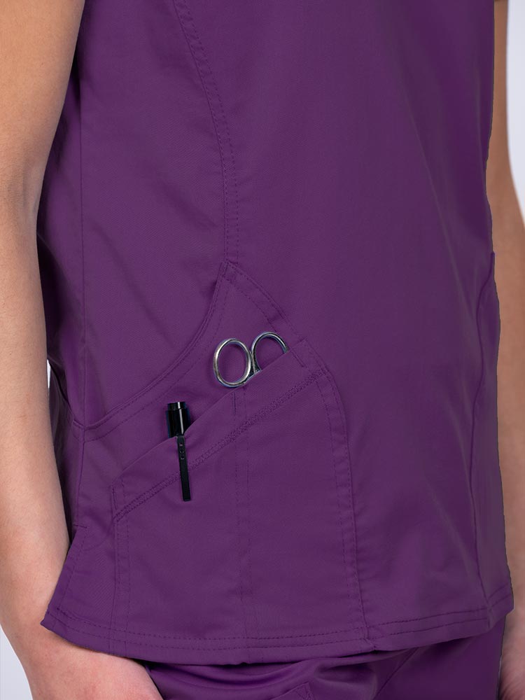 Woman wearing an Epic by MedWorks Women's Knit Collar Mock Wrap Scrub Top in eggplant with two outside utility pockets on the wearer's right side.