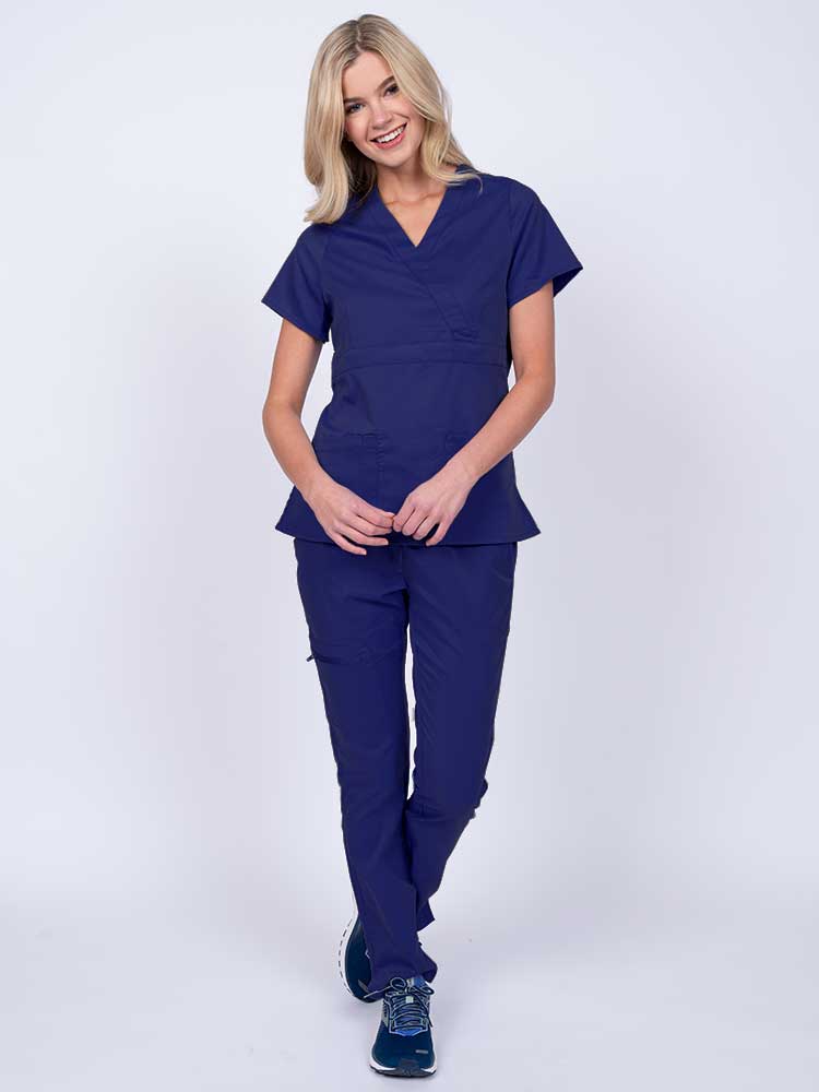 A young female healthcare worker wearing an Epic by MedWorks Women's Mock Wrap Scrub Top in navy featuring a super soft, 2-way stretch fabric.