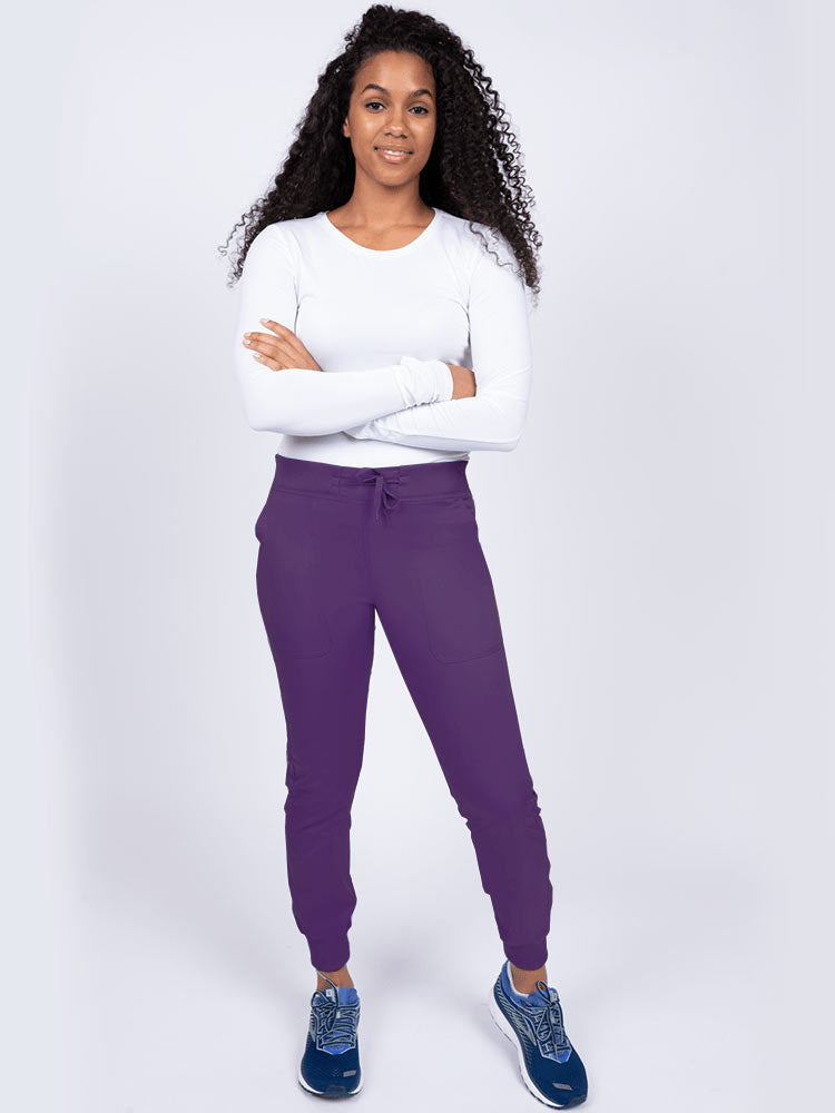 A young lady nurse wearing an Epic by MedWorks Women's Yoga Jogger Scrub Pant in Eggplant size Large Petite featuring stylish cover stitch detail throughout.