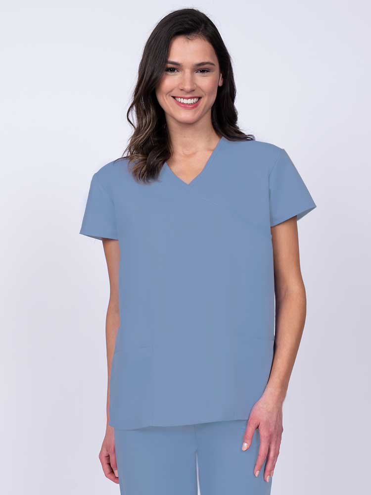 Young woman wearing a Luv Scrubs by MedWorks Women's Mock Wrap Scrub Top in ceil featuring a Y-neckline and side slits for additional range of motion.