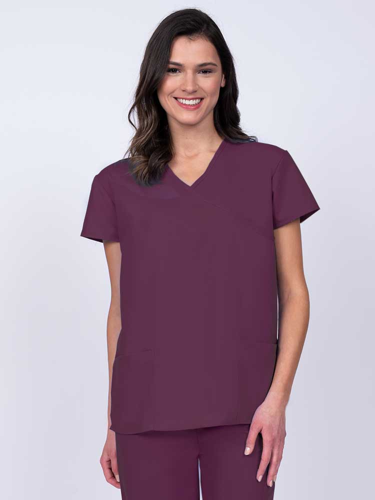 Young woman wearing a Luv Scrubs by MedWorks Women's Mock Wrap Scrub Top in wine featuring a Y-neckline and side slits for additional range of motion.