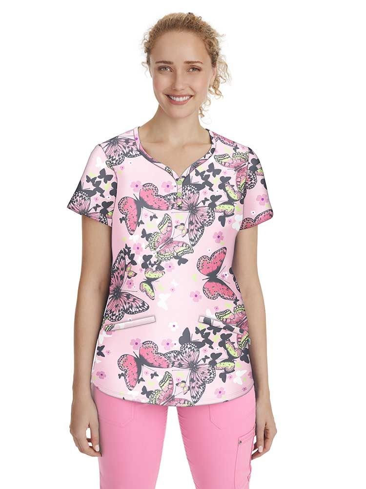 A young female Pediatric Nurse wearing a Women's Isabel Printed Scrub Top in "Butterfly's Journey" featuring a y-neck and two front angled pockets.