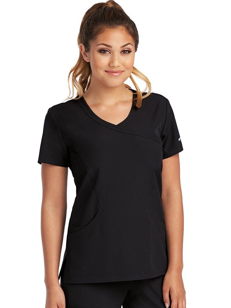 A young female Nurse Practitioner wearing a Skechers Women's Reliance Mock Wrap Scrub Top in Black size small featuring a crossover mock wrap neckline with shoulder yokes to provide a flattering all day fit.