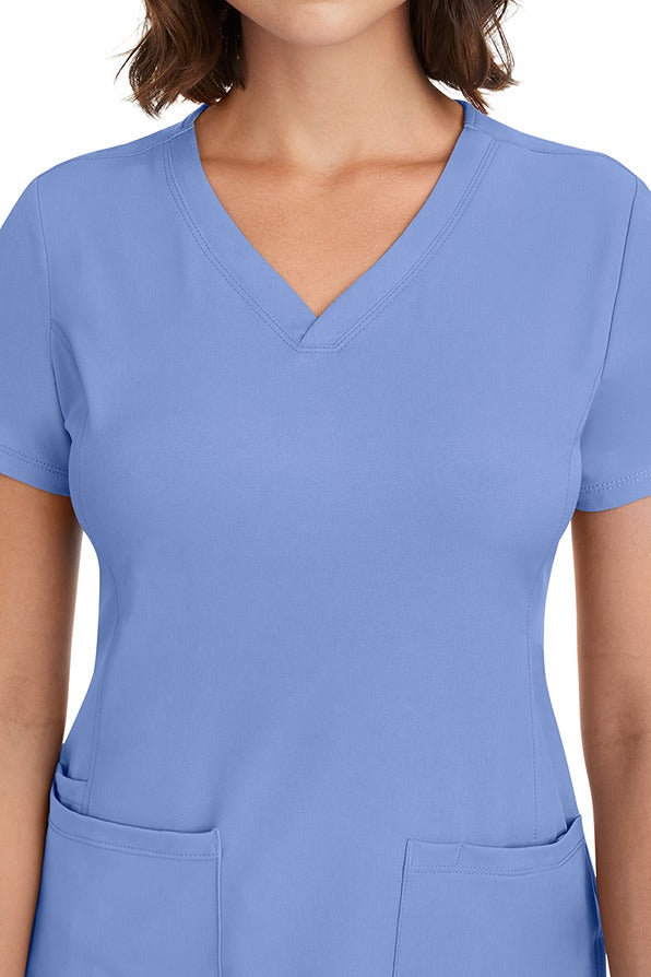 A young woman CNA wearing a HH-Works Women's Monica Multi-Pocket Scrub Top in Ceil featuring front princess seaming to ensure a flattering fit.