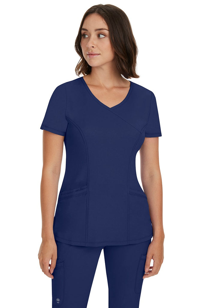 A young Home Care Registered Nurse wearing a HH-Works Women's Madison Mock Wrap Scrub Top in Navy featuring a total of 4 pockets for all of your on the go storage needs.