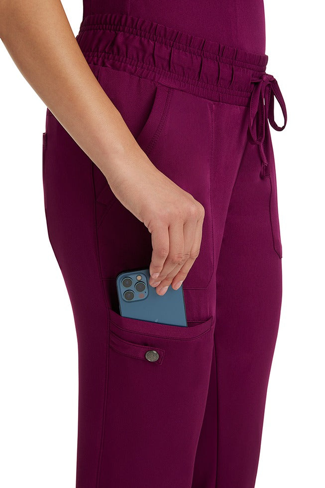 A female LPN wearing an HH Works Women's Renee Jogger Scrub Pant in Wine featuring 2 front patch pockets & 1 cargo pocket the wearer's right side leg.