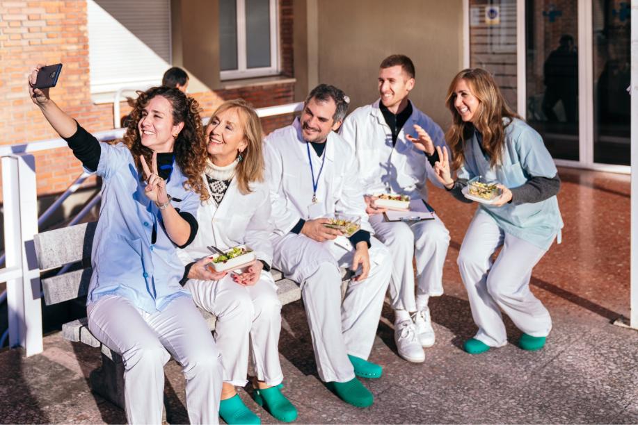 Group of happy healthcare professionals in scrubs taking a selfie and enjoying a meal break outdoors. Workplace camaraderie and self-care in healthcare.