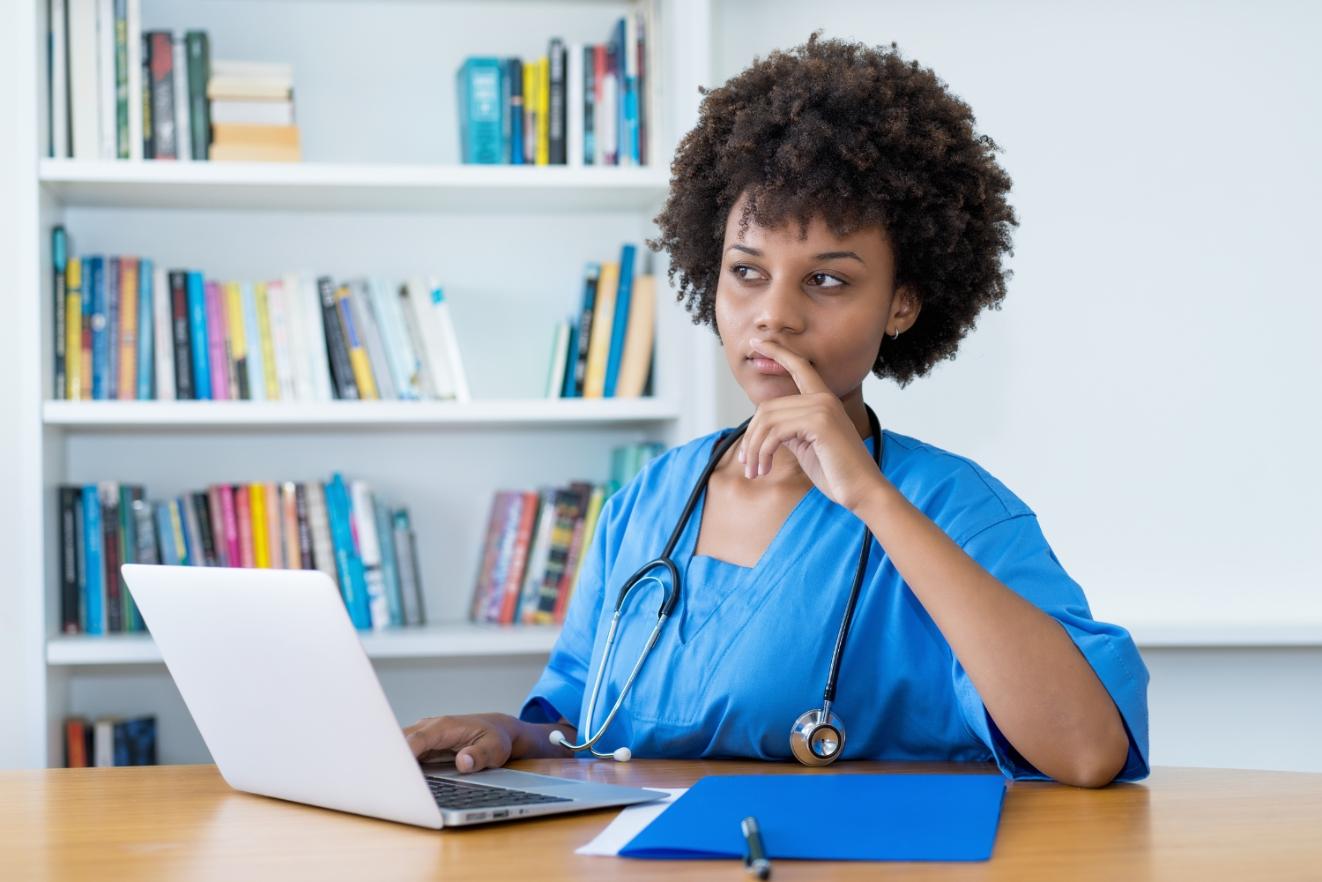 Nurse in blue scrubs at a desk, thinking about changing specialties while working on a laptop, with books in the background.