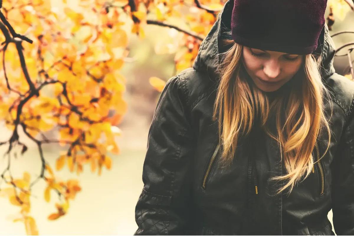 Sad Woman wearing a jacket and hat in the fall, with leaves in the background.