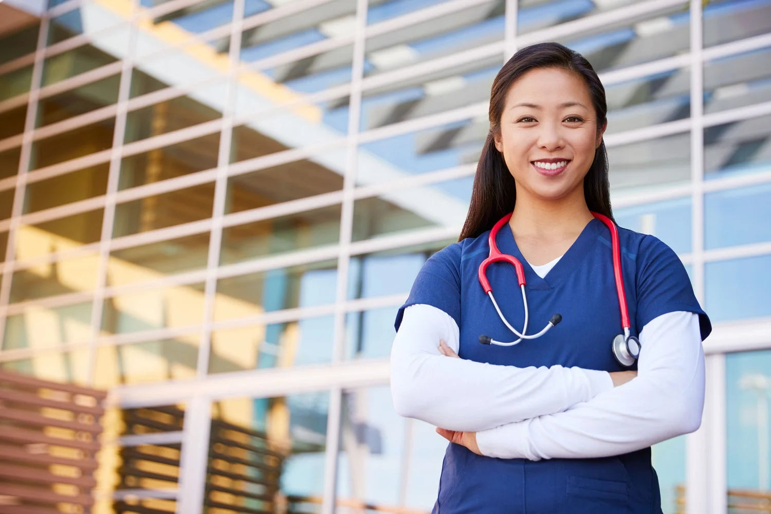 A young female Surgical Assistant wearing Navy Blue scrubs with a White long sleeve underscrub and a red stethoscope, outside of a hospital.