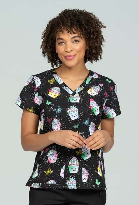 A young female Medical Assistant wearing a Meraki Sport Women's Print Scrub Top in "Cake It Easy" size 3XL featuring a v-neckline & short sleeves.