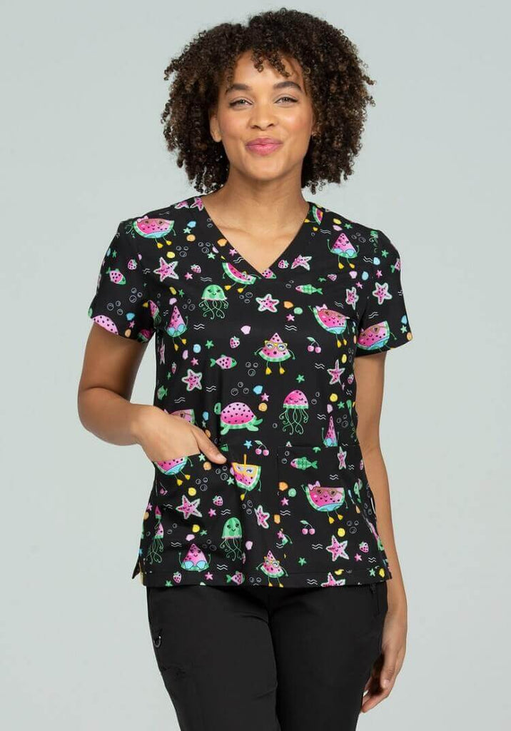 A young female Pediatrician wearing a Large Meraki Sport Women's Print Scrub Top in "Stuck on You" size Medium featuring a v-neckline & short sleeves.