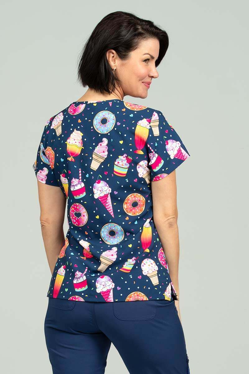 A young female Medical Assistant wearing a Meraki Sport Women's Print Scrub Top in "Sweets & Treats" featuring side slits for additional range of motion & shoulder yokes for shaping.