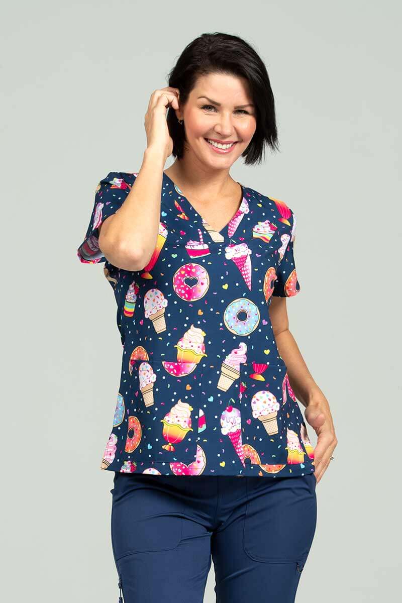 A female Nursing Assistant wearing a Meraki Sport Women's Print Scrub Top in "Sweets & Treats" size Large featuring a contemporary fit with a V-neckline.