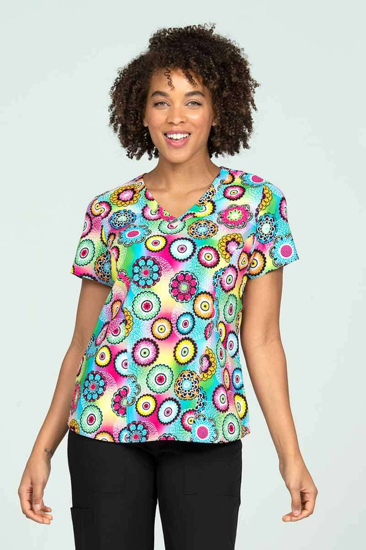 A young female Pediatrician wearing a Meraki Sport Women's Print Scrub Top in "Pop of Fun" size Medium featuring 2 front patch patch pockets & a V-neckline.