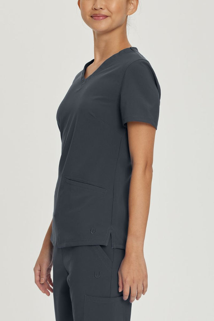 A young female Nursing Assistant wearing an Urbane Performance Women's Motivate V-neck Scrub Top in Graphite size XS featuring side slits with mesh detail to provide additional range of motion and added comfort.