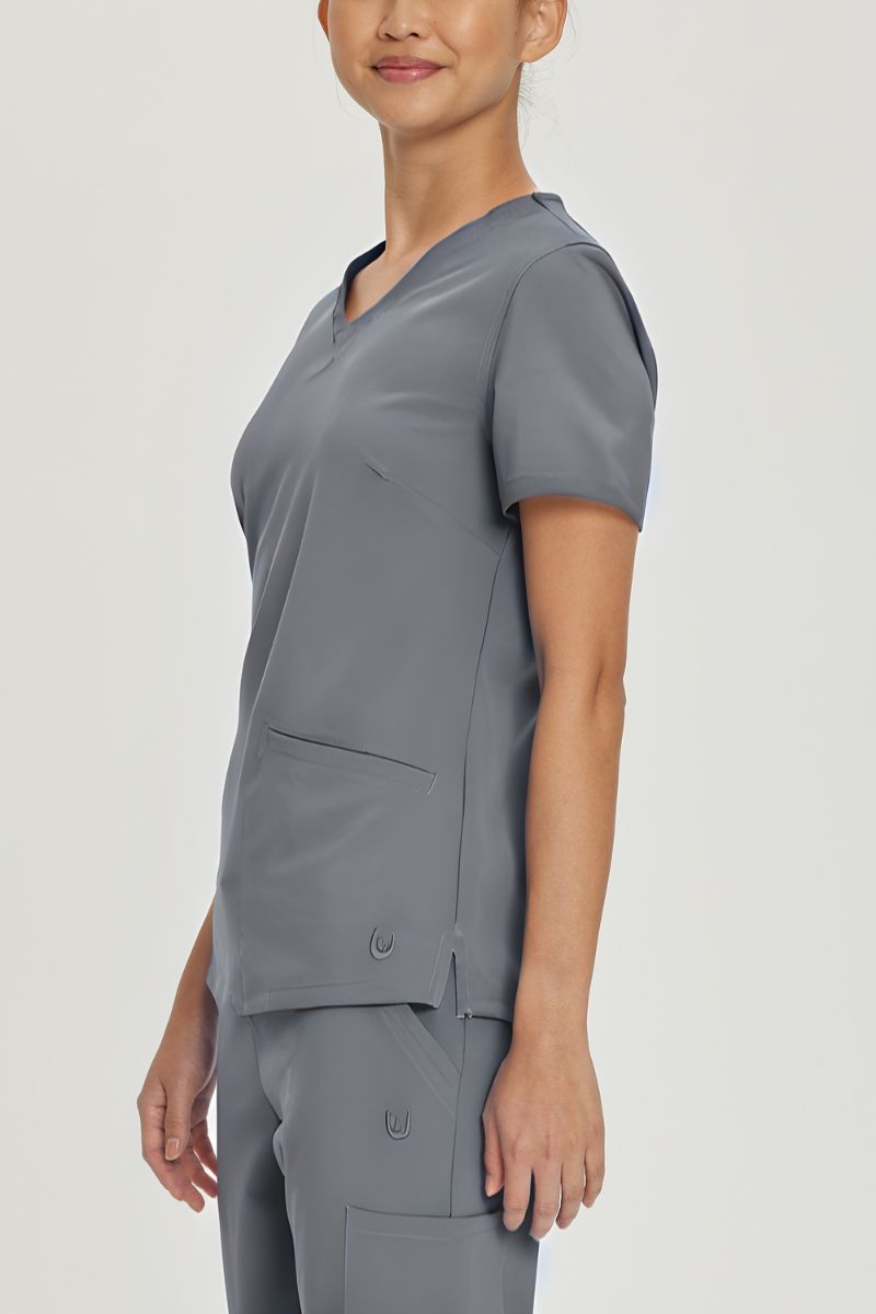 A young female Nursing Assistant wearing an Urbane Performance Women's Motivate V-neck Scrub Top in Steel Grey size XS featuring side slits with mesh detail to provide additional range of motion and added comfort.