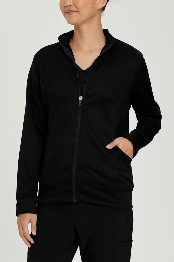 A young female LPN wearing an Urbane Performance Women's Zip-Up Scrub Jacket in Black size Medium featuring three pockets, including a hidden pocket with a zipper closure.