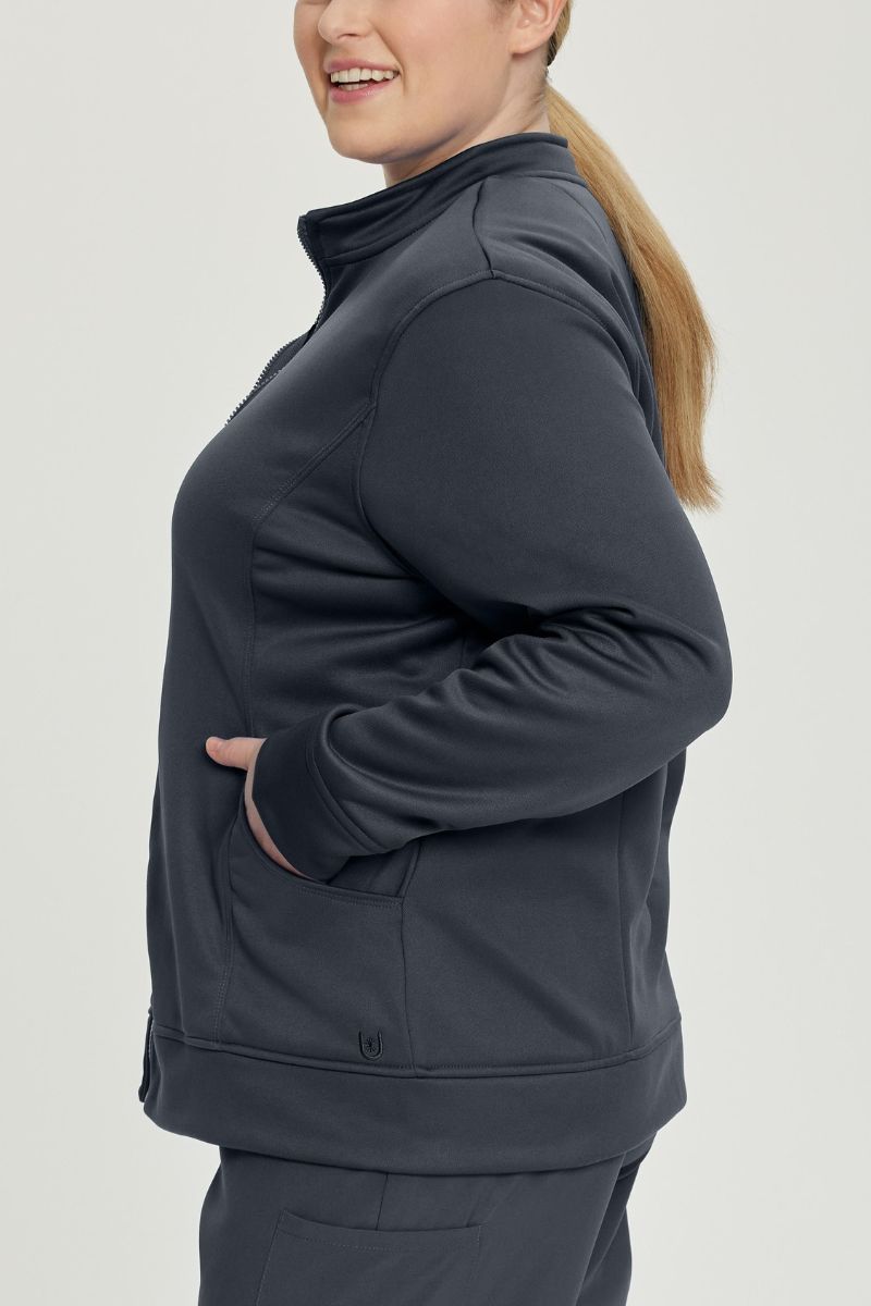 A young female Nurse wearing an Urbane Performance Women's Zip-Up Scrub Jacket in Graphite featuring three pockets, including a hidden pocket with a zipper closure.