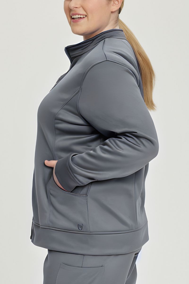 A young female Nursing Assistant wearing an Urbane Performance Women's Zip-Up Scrub Jacket in Steel featuring three pockets, including a hidden pocket with a zipper closure.