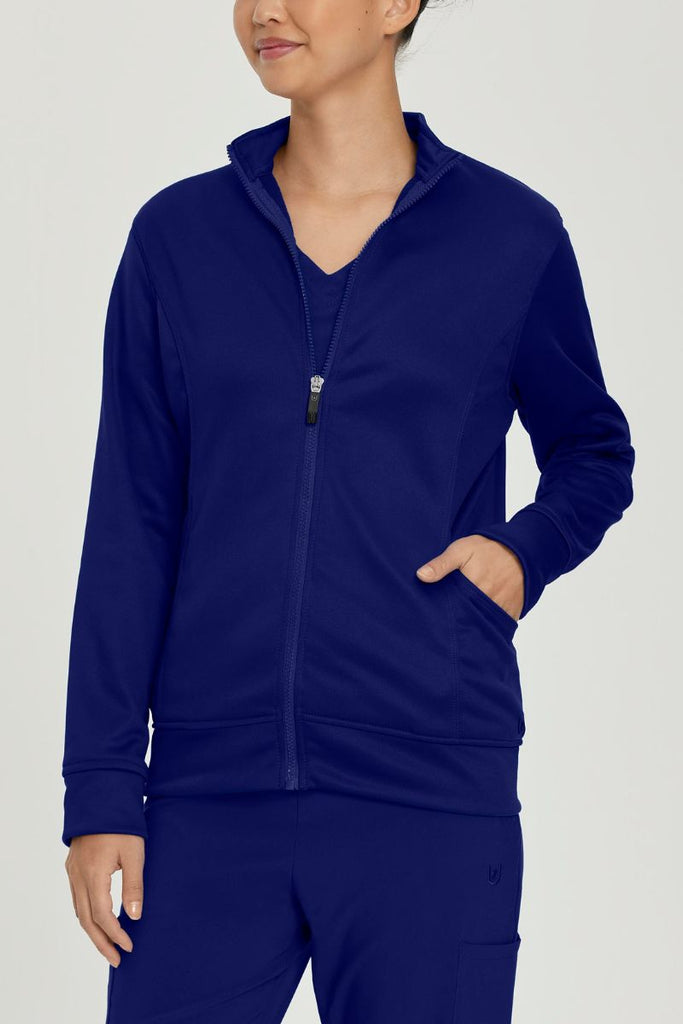A young female Nurse wearing an Urbane Performance Women's Zip-Up Scrub Jacket in True Navy size XS featuring three pockets, including a hidden pocket with a zipper closure.