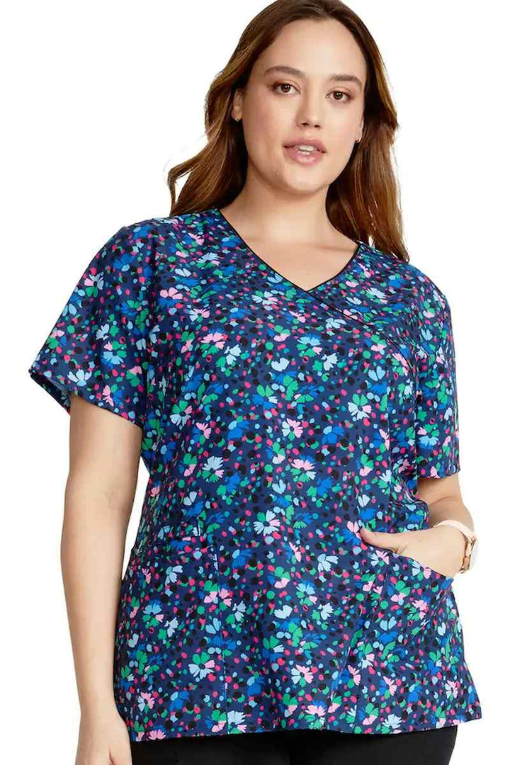 A young female Pediatric Nurse wearing a Cherokee Women's Mock Wrap Print Scrub Top in Confetti Daisies featuring a modern classic fit.