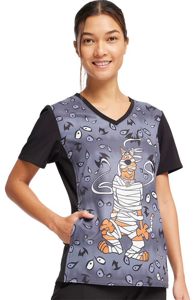 A young female Pediatric Nurse wearing a Tooniforms Women's V-Neck Printed scrub Top in "Under Wraps" featuring Scooby Doo dressed up as a mummy on a light grey background with creepy eyes and bats scattered throughout.