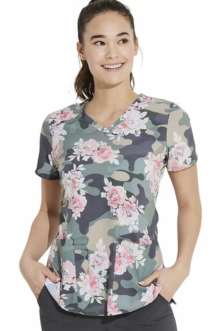 A young female nurse wearing a Dickies Women's V-Neck Printed Scrub Top in Camo's Abloom featuring two spacious front pockets for on the go storage needs. 