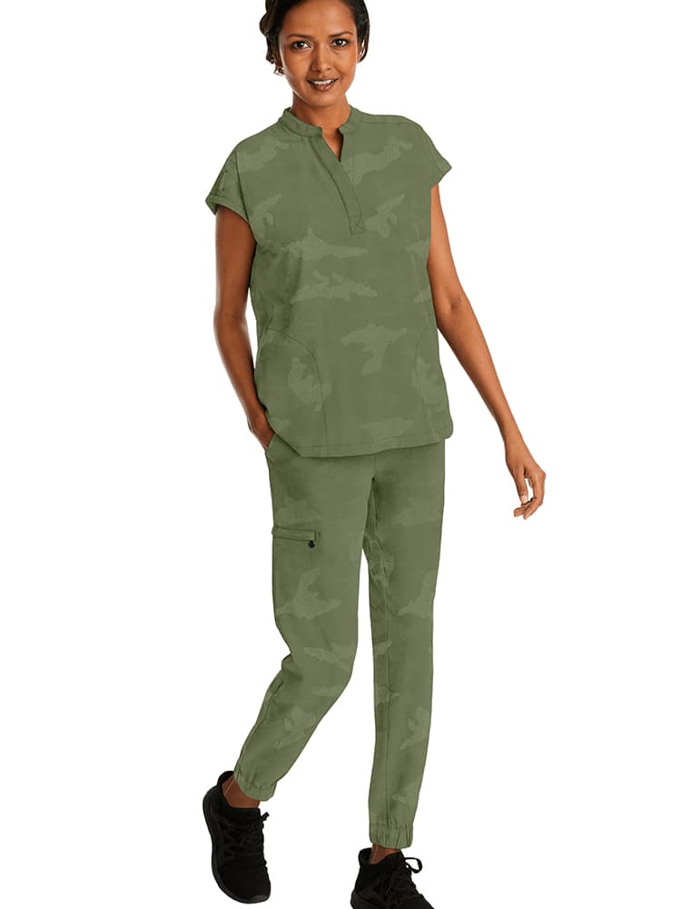 A young female Surgical Assistant wearing a Purple Label Women's Journey Camo Scrub Top in Olive size Medium featuring a modern fit and Mandarin one button collar.