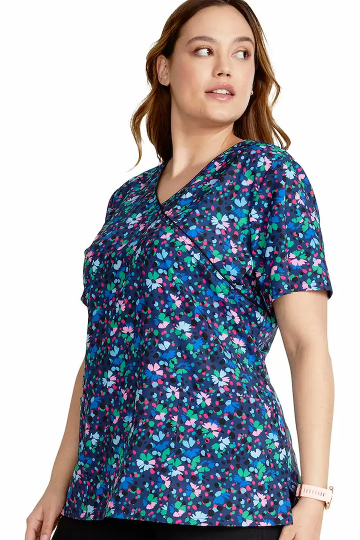 A young female nurse wearing a Cherokee Women's Mock Wrap Print Scrub Top in Confetti Daisies featuring side vents to enhance freedom of movement and provide a comfortable all-day fit.