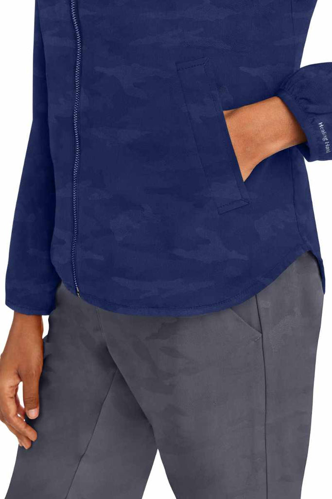 A young female LPN wearing a Purple Label Women's Destini Camo Scrub Jacket in Navy size Medium featuring 2 front angled pockets.